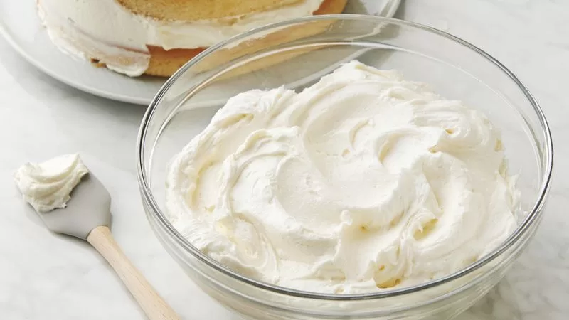 Un tazón de vidrio lleno con betún de queso crema extra firme, con una espatula pequeña sobre una mesa.